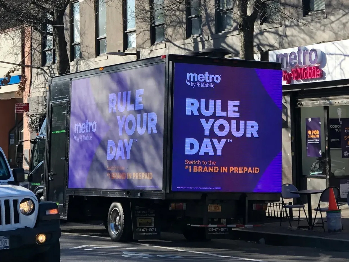 Metro by T-Mobile advertisement on a truck displaying "RULE YOUR DAY" and promoting the "#1 BRAND IN PREPAID" services, set in an urban environment.